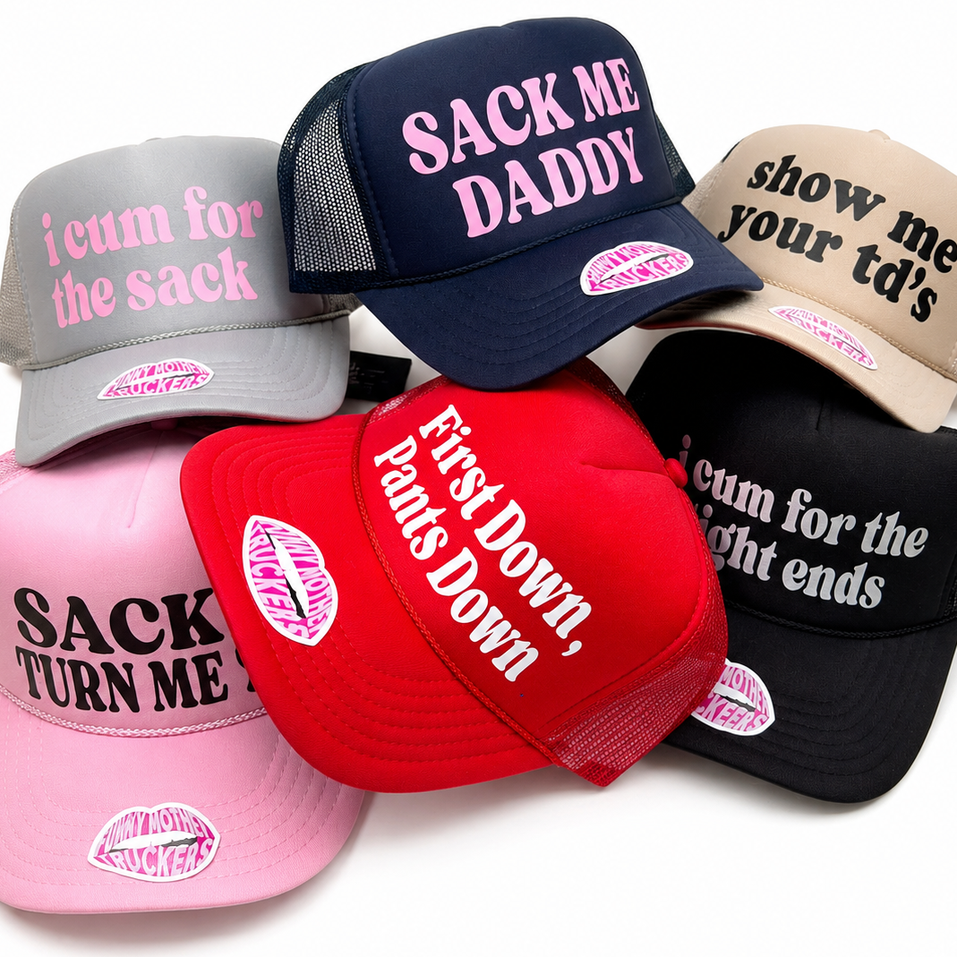 Six trucker-style caps in different colors are arranged in a pile on a white background. The hats are in gray, navy, beige, pink, red, and black, and each has bold text on the front panel with suggestive phrases like 'SACK ME DADDY' and 'First Down, Pants Down.