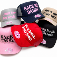 Six trucker-style caps in different colors are arranged in a pile on a white background. The hats are in gray, navy, beige, pink, red, and black, and each has bold text on the front panel with suggestive phrases like 'SACK ME DADDY' and 'First Down, Pants Down.