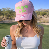 Woman wearing a pink cap with text, holding a can on a golf course.