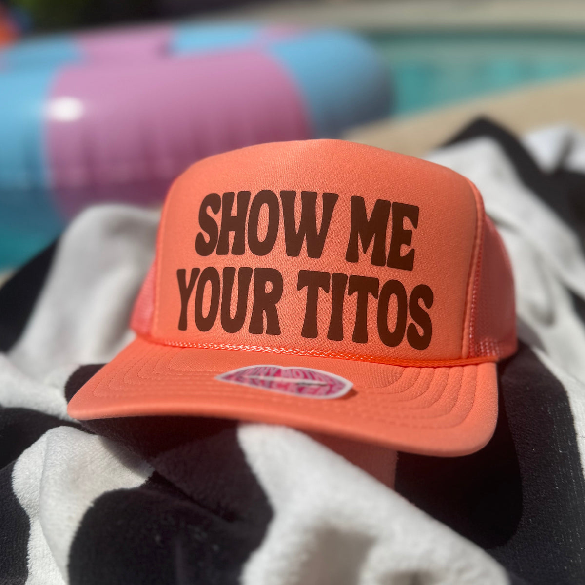 Orange cap with 'SHOW ME YOUR TITOS' text on a towel by a pool.