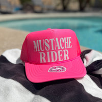 Pink cap with 'Mustache Rider' text on a striped towel by a pool