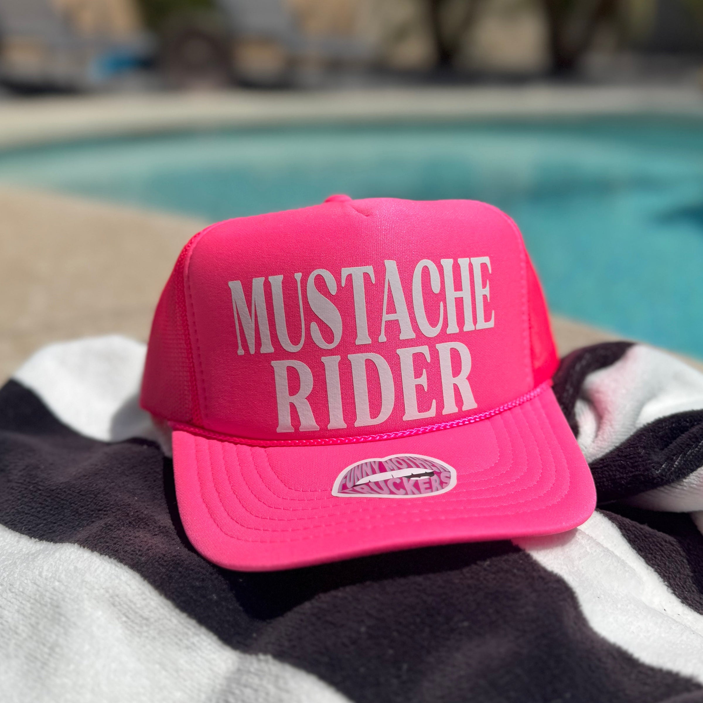 Pink cap with 'Mustache Rider' text on a striped towel by a pool