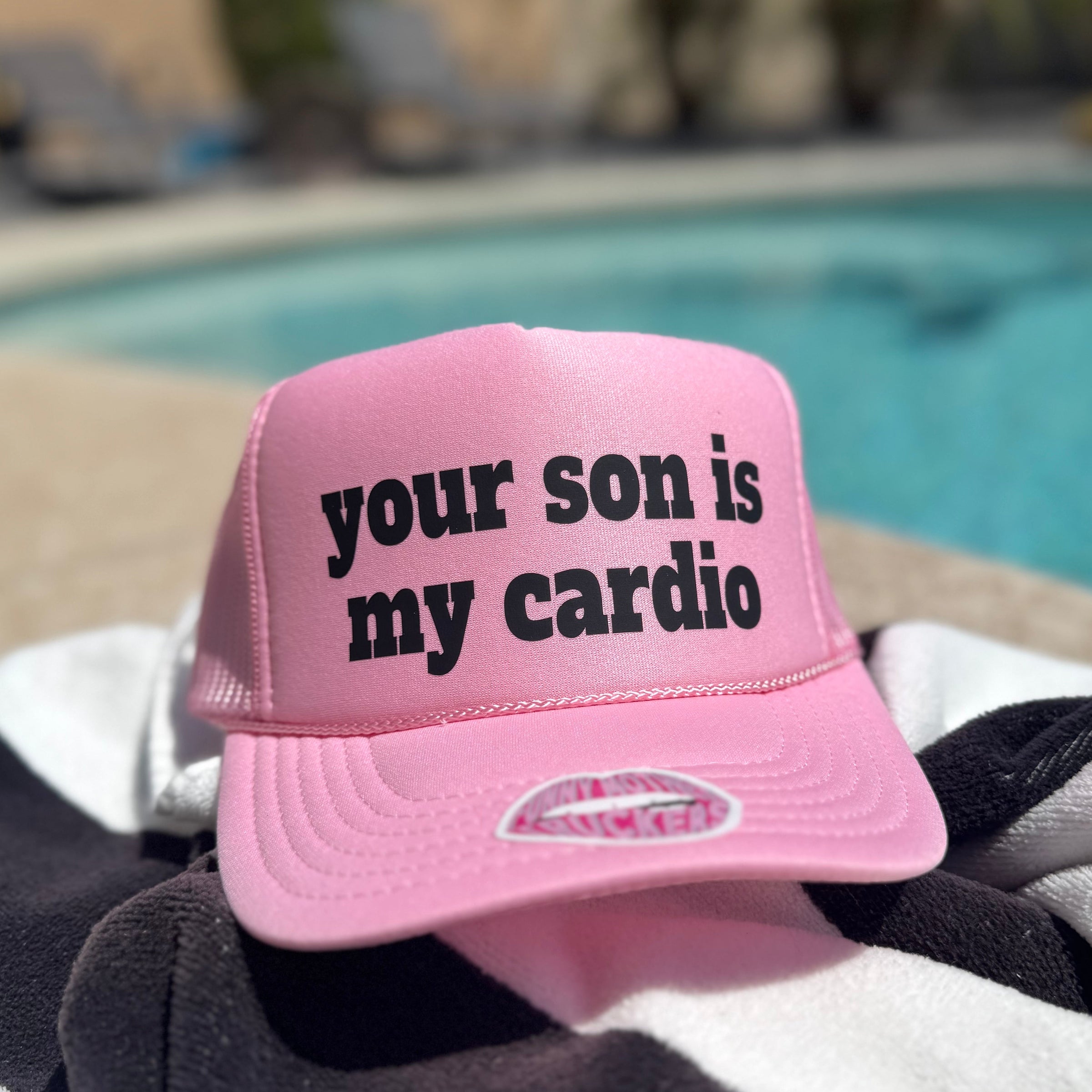 Pink cap with text 'your son is my cardio' on a towel by a pool.