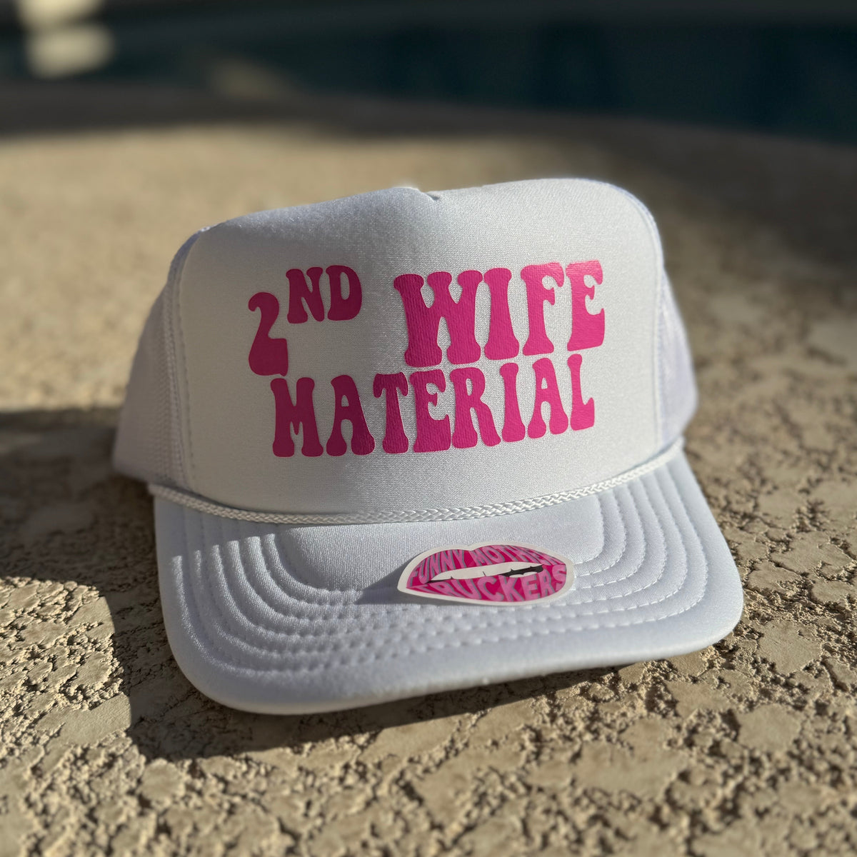 White cap with pink text '2nd Wife Material' on a sandy surface.
