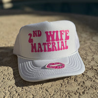 White cap with pink text '2nd Wife Material' on a sandy surface.