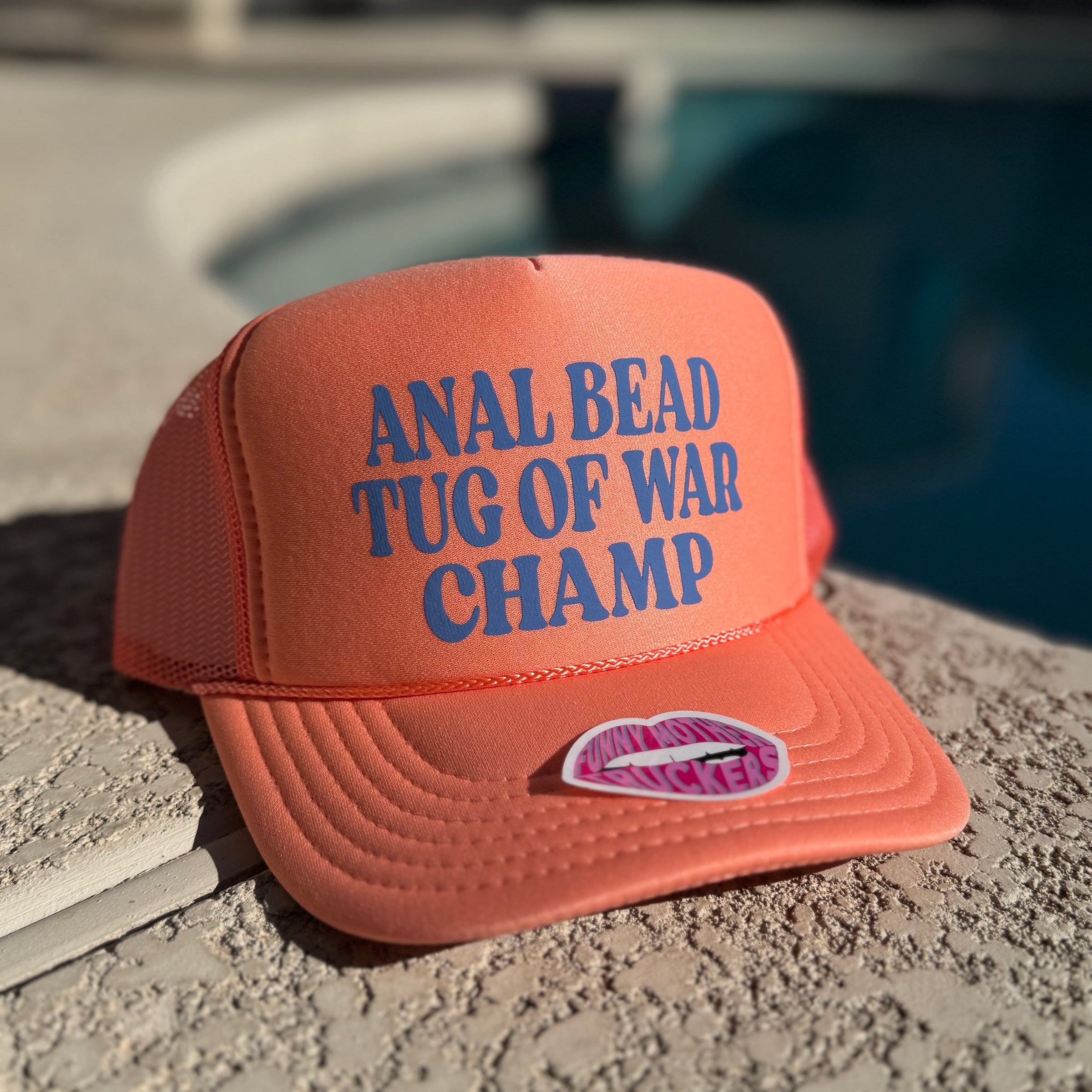 Orange cap with blue text 'Anal Bead Tug of War Champ' on a concrete surface.