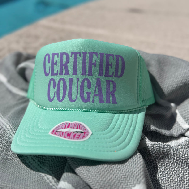Mint green cap with 'CERTIFIED COUGAR' text and pink lips graphic on a gray surface.