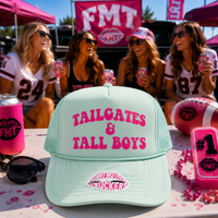A mint green trucker hat with pink text reading 'TAILGATES & TALL BOYS' and a lip sticker on the brim, displayed on a table with pink tailgate-themed props and four women sitting in the background at an outdoor event.