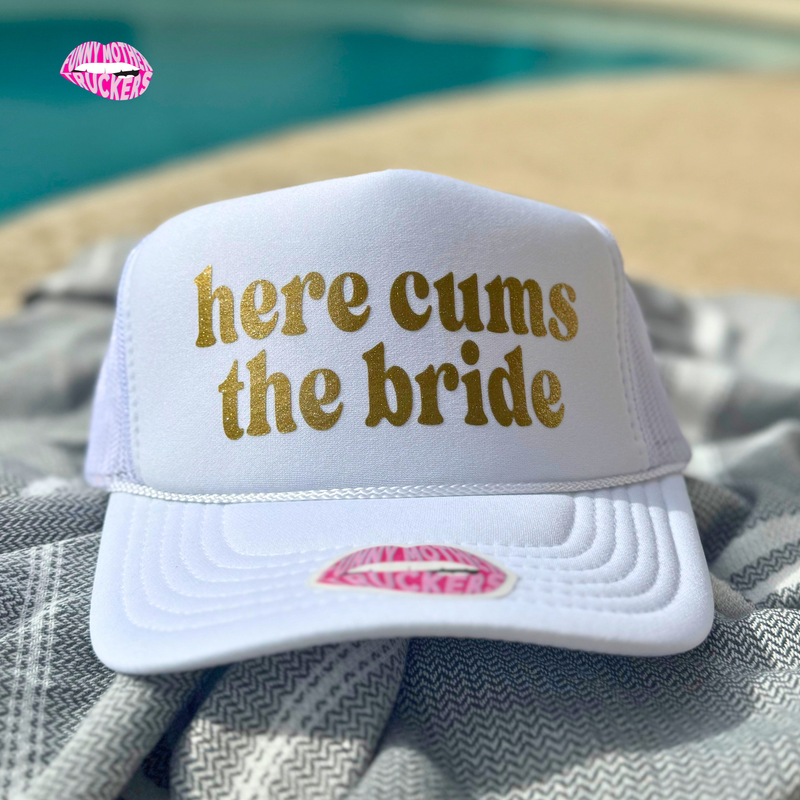 White cap with 'here cums the bride' text on a towel by a pool.