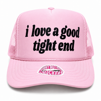 A light pink trucker hat with black embroidered text reading 'i love a good tight end' on the front panel and a pink and white 'Jelly Hornets Buckers' patch on the brim, shown against a plain white background.