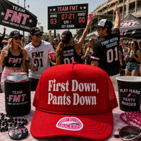 A red trucker hat with white text reading 'First Down, Pants Down' is displayed on a table in a lively outdoor sports event setting. The scene features people wearing matching black 'Team FMT Funny Mother Truckers' shirts and hats, with a scoreboard and branded cups, cans, and beads in the background.