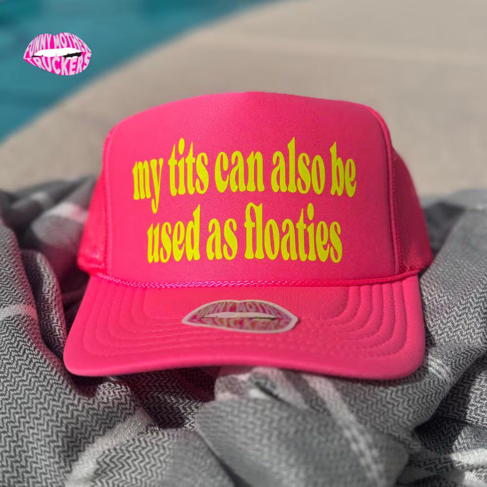 Pink cap with yellow text 'my tits can also be used as floaties' on a towel by a pool.