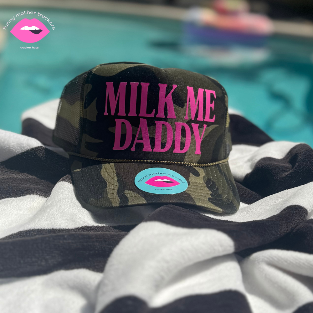 A camouflaged trucker hat with 'Milk Me Daddy' written in pink on the front, placed on a towel beside a pool.