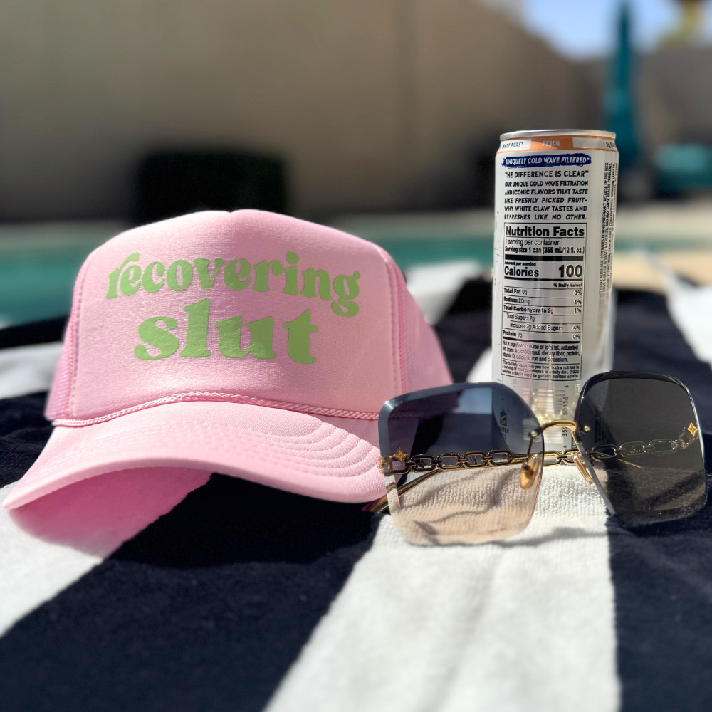 A pink trucker hat with the text 'recovering slut' written in green letters, displayed next to a can of beer and a pair of sunglasses, placed on a striped surface.
