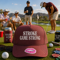 A maroon trucker-style cap with white text reading 'STROKE GAME STRONG' and a pink lips patch that says 'STROKED BACK' is placed on a golf course. Four men are visible in the background, with one actively swinging a golf club.
