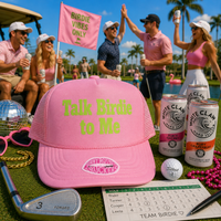 A pink trucker hat with 'Talk Birdie to Me' in neon yellow-green text and a lip print on the brim, displayed on a golf course with golf clubs, cans of White Claw alcohol-free canned beer, a golf ball, a scoreboard, and a pink flag in the background with people celebrating.